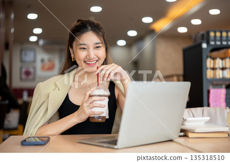 Asian woman enjoy drinking iced tea or coffee while looking at a laptop on wooden table in the cafe. Asian woman enjoy drinking iced tea or coffee while looking at a laptop on wooden table in the cafe. 135318510