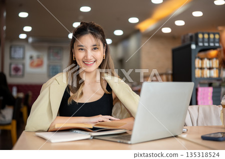 Smiling asian woman folding arms over notebook aside laptop while sitting at wooden table in a cafe. 135318524