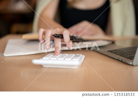 Close up woman's hand holding a pen and using calculator aside notebook and laptop on wooden table. Close up woman's hand holding a pen and using calculator aside notebook and laptop on wooden table. 135318533