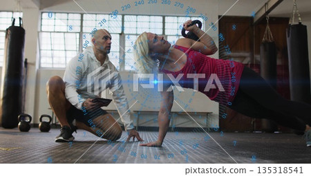 Performing side plank row with kettlebell fitness client in studio gym, trainer guiding on tablet 135318541