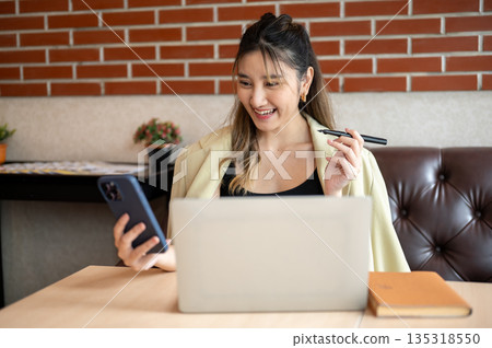 Asian woman holding pen and looking at phone in front of a laptop on table while sit on sofa in cafe Asian woman holding pen and looking at phone in front of a laptop on table while sit on sofa in cafe 135318550