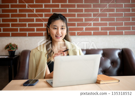 Asian woman thinking as she holding pen pointing at laptop on table while sitting on sofa in a cafe. 135318552