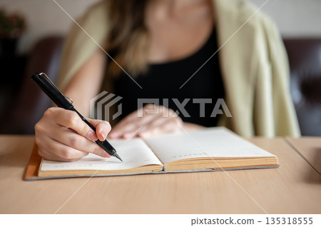 Close up woman's hand holding fountain pen writing in notebook on table while sits on sofa in a cafe 135318555