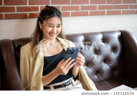 Smiling asian woman holding credit card and looking at phone while sitting on leather sofa in cafe. 135318559