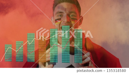 Shouting sports fan holding soccer ball in studio, with red cape, flag face paint, data charts 135318580