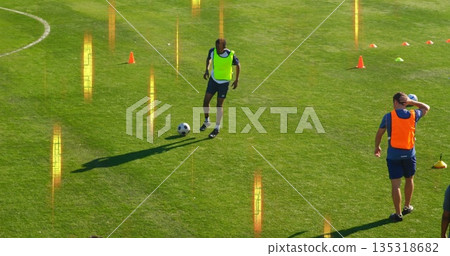 Dribbling yellow bib athlete controlling soccer ball on grass pitch, with orange and green cones Dribbling yellow bib athlete controlling soccer ball on grass pitch, with orange and green cones 135318682