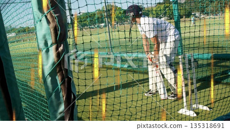 Practicing male cricket batsman in white uniform holding bat at net cage on field, with stumps 135318691