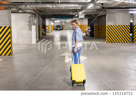 A woman smiles as she pulls a yellow suitcase through an underground parking garage A woman smiles as she pulls a yellow suitcase through an underground parking garage 135318752