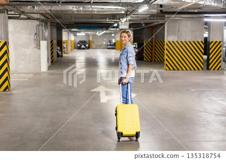 A smiling woman stands in a parking garage with a yellow suitcase, ready for travel A smiling woman stands in a parking garage with a yellow suitcase, ready for travel 135318754