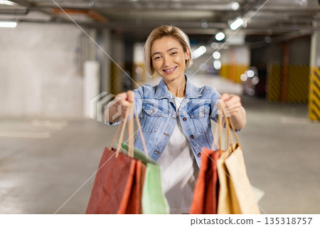 A smiling woman holds shopping bags in a parking garage, ready to share her purchases 135318757