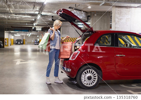 A woman smiles while putting shopping bags into the trunk of her red car in a parking garage A woman smiles while putting shopping bags into the trunk of her red car in a parking garage 135318760