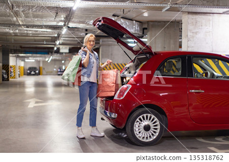 A smiling woman loads shopping bags into the trunk of her red car in a parking garage A smiling woman loads shopping bags into the trunk of her red car in a parking garage 135318762