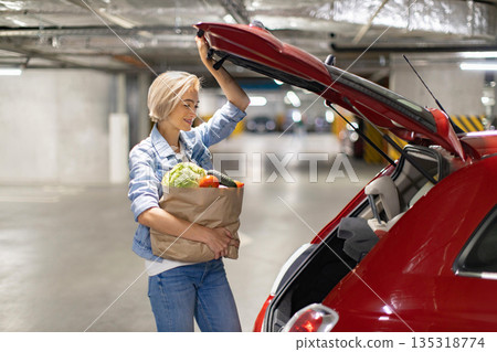 A woman smiles while loading groceries into the trunk of her car in a parking garage A woman smiles while loading groceries into the trunk of her car in a parking garage 135318774