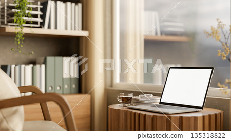 White screen laptop and coffee on tree stump table with armchair and bookshelf in sunlit living room 135318822