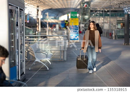 Young woman walking through a bright modern airport terminal with suitcase, moving toward departure or arrival in sleek glass-and-metal architecture, solo traveler on the go Young woman walking through a bright modern airport terminal with suitcase, moving toward departure or arrival in sleek glass-and-metal architecture, solo traveler on the go 135318915