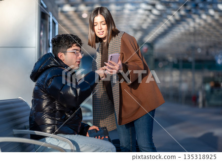 Young couple standing and sitting in a modern urban train station, looking at an electronic device and having a conversation about content on smartphone, sharing a lifestyle moment Young couple standing and sitting in a modern urban train station, looking at an electronic device and having a conversation about content on smartphone, sharing a lifestyle moment 135318925