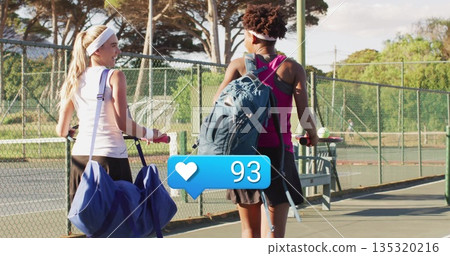 Walking two tennis players sporting gear carrying rackets and bags on sunlit court, with ball cart 135320216