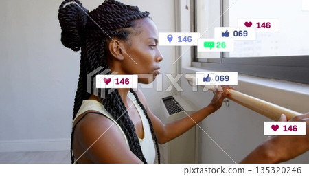 Looking dancer holding barre in tank top by window in studio, with floating reaction icons 135320246