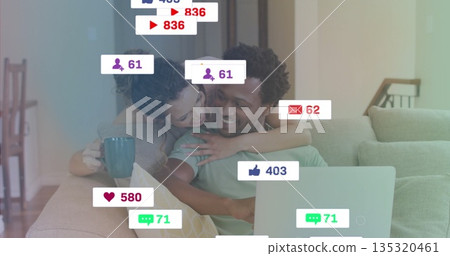 Smiling couple hugging on couch, using laptop browsing with floating icons, holding teal coffee mug 135320461
