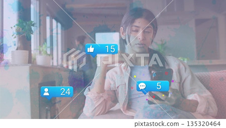 Teenage girl wearing pink overshirt checking smartphone in shared lounge, with laptop, plants 135320464