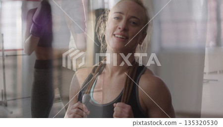Smiling athletic woman holding jump rope over shoulders in gym, showing boxing gloves reflection Smiling athletic woman holding jump rope over shoulders in gym, showing boxing gloves reflection 135320530