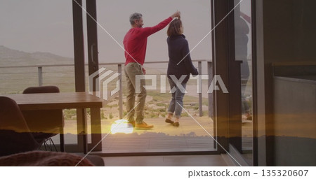 Spinning couple wearing sweaters dancing on deck outside sliding glass doors, overlooking valley 135320607