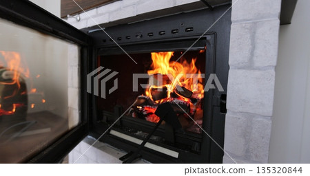 Stirring wood with poker in the fireplace. Warm cozy burning fire in a brick fireplace close. 135320844