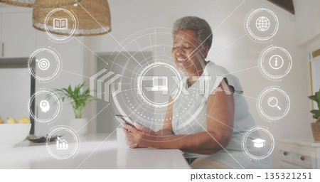 Smiling senior woman leaning on white island in home kitchen, holding smartphone with network icons Smiling senior woman leaning on white island in home kitchen, holding smartphone with network icons 135321251