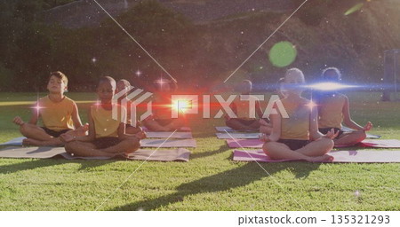 Meditating children sitting cross-legged on yoga mats in park on grassy lawn, wearing yellow tops 135321293