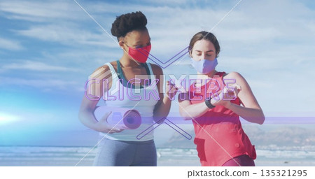 Standing women wearing sportswear and face masks on beach, holding yoga mat and checking smartwatch 135321295