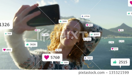 Posing woman wearing sweater holding smartphone near lakeside, with floating social media icons Posing woman wearing sweater holding smartphone near lakeside, with floating social media icons 135321599