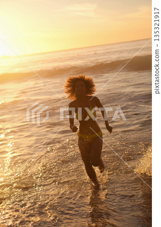 African American woman running through ocean waves on beach splashing water in yellow bikini African American woman running through ocean waves on beach splashing water in yellow bikini 135321917