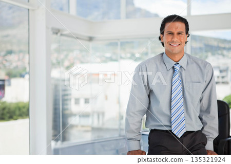 Mid adult man sitting at desk near floor-to-ceiling windows in modern office workspace, copy space 135321924