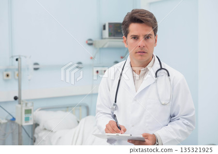 Male doctor in lab coat holding clipboard and stethoscope, checking vital signs monitor at hospital Male doctor in lab coat holding clipboard and stethoscope, checking vital signs monitor at hospital 135321925