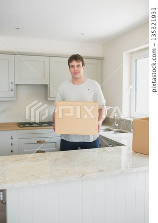 Middle-aged man holding cardboard box behind marble home kitchen island under natural window light 135321931