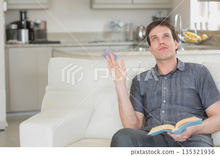 Mid adult man reading paperback and tossing smartphone on white sofa in lounge-kitchen, copy space 135321936