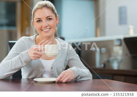 Woman holding ceramic coffee mug and saucer while smiling at kitchen table with coffee maker 135321938