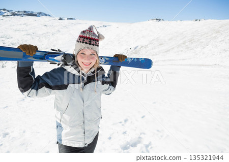 Woman in twenties holding pair of blue skis across shoulders on snowy mountain slope, copy space 135321944