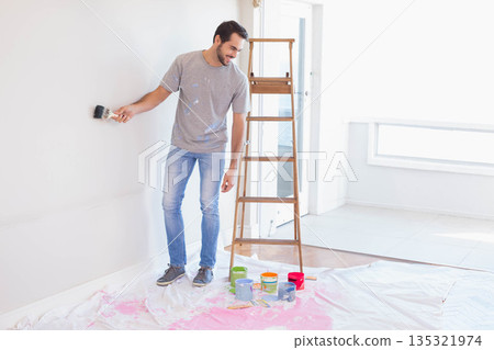 Male homeowner painting wall inside empty room, holding paintbrush near step ladder with paint cans Male homeowner painting wall inside empty room, holding paintbrush near step ladder with paint cans 135321974