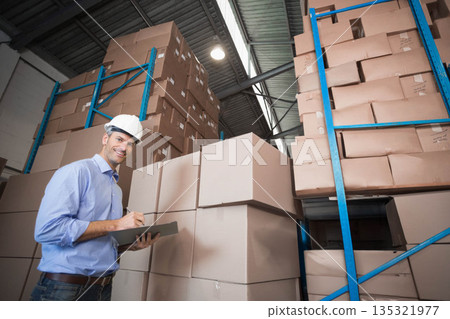 Middle aged male wearing hard hat in warehouse inspecting clipboard among boxes and blue racks Middle aged male wearing hard hat in warehouse inspecting clipboard among boxes and blue racks 135321977