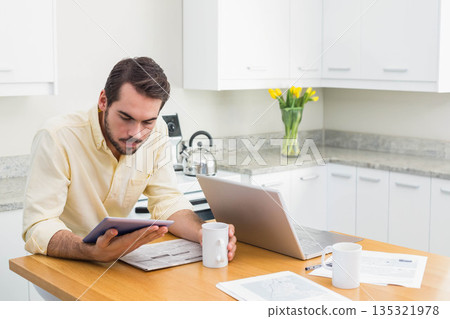 Man working on tablet while holding ceramic coffee mug at kitchen island with laptop and documents 135321978