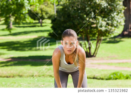 Woman pausing leaning forward with hands on knees in sunlit grassy park wearing sportswear Woman pausing leaning forward with hands on knees in sunlit grassy park wearing sportswear 135321982