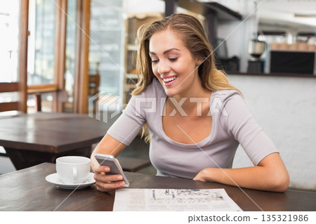 Woman sitting at wooden table inside cafe wearing gray top smiling at smartphone beside coffee cup Woman sitting at wooden table inside cafe wearing gray top smiling at smartphone beside coffee cup 135321986