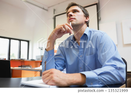 Man sitting in meeting room at table holding pen near chin, viewing projection screen and notebook Man sitting in meeting room at table holding pen near chin, viewing projection screen and notebook 135321999