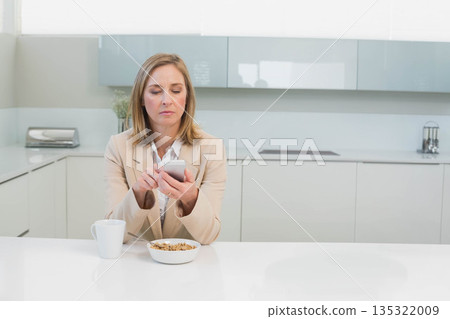 Senior woman sitting at modern kitchen island holding smartphone beside cereal bowl with milk 135322009
