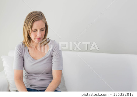 Senior woman sitting on white sofa in living room touching cushion and looking downward, copy space 135322012