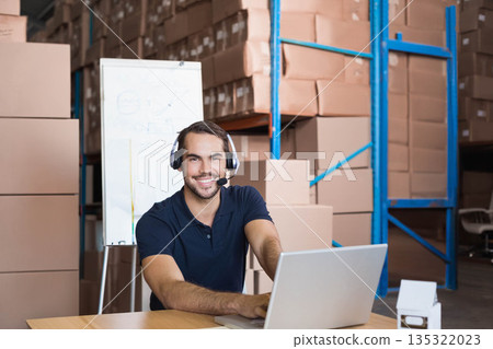 Man wearing polo with headset mic managing inventory on laptop at warehouse table with flip chart Man wearing polo with headset mic managing inventory on laptop at warehouse table with flip chart 135322023