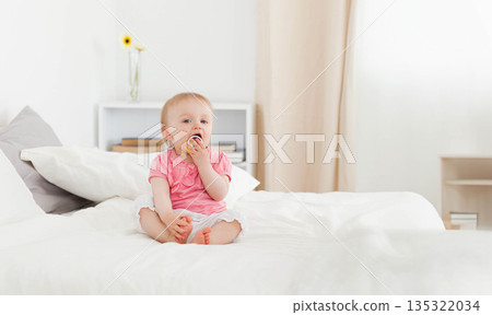 Female infant eating snack while sitting on white bed near vase with yellow flower and books Female infant eating snack while sitting on white bed near vase with yellow flower and books 135322034