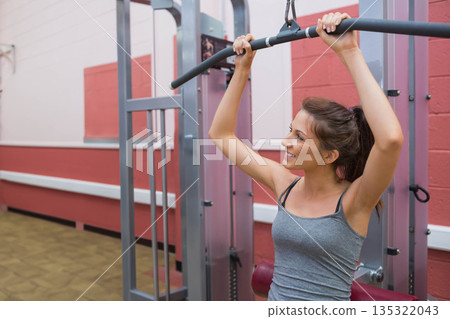 Female gym member pulling curved bar downward on lat pulldown machine in weight area, copy space Female gym member pulling curved bar downward on lat pulldown machine in weight area, copy space 135322043