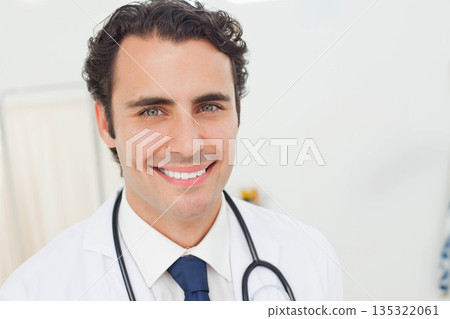 Male doctor wearing white lab coat and stethoscope, smiling in exam room with shelves Male doctor wearing white lab coat and stethoscope, smiling in exam room with shelves 135322061